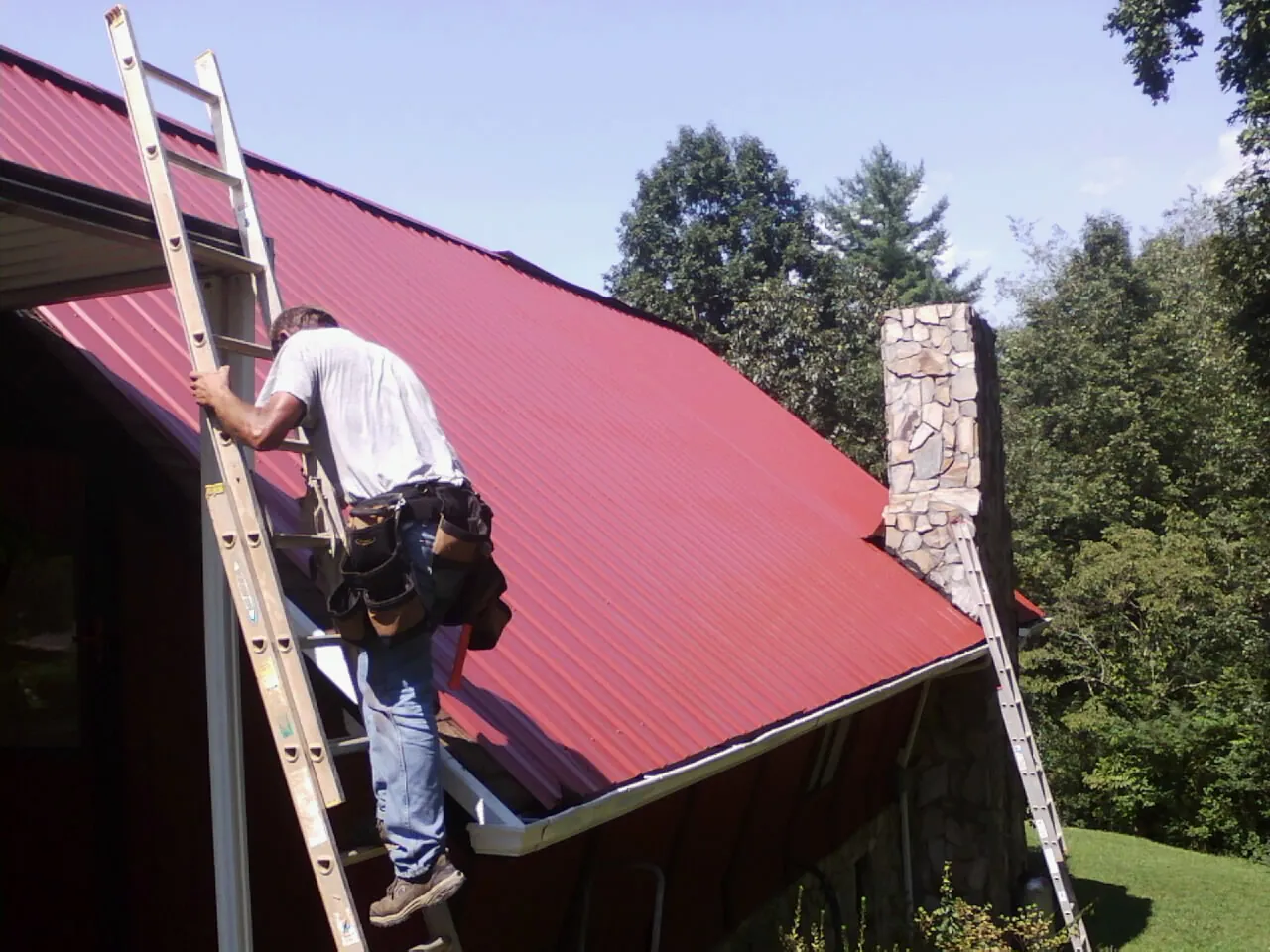 Roofer on a ladder beside a red metal roof and stone chimney — Soffit & Fascia Repair by Roofing Brothers in Daleville
